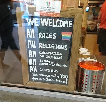 Placa colocada num café e escrita em inglês que dá boas-vindas a pessoas de qualquer gênero e qualquer nacionalidade, diz que todos podem se sentir seguros neste estabelecimento
